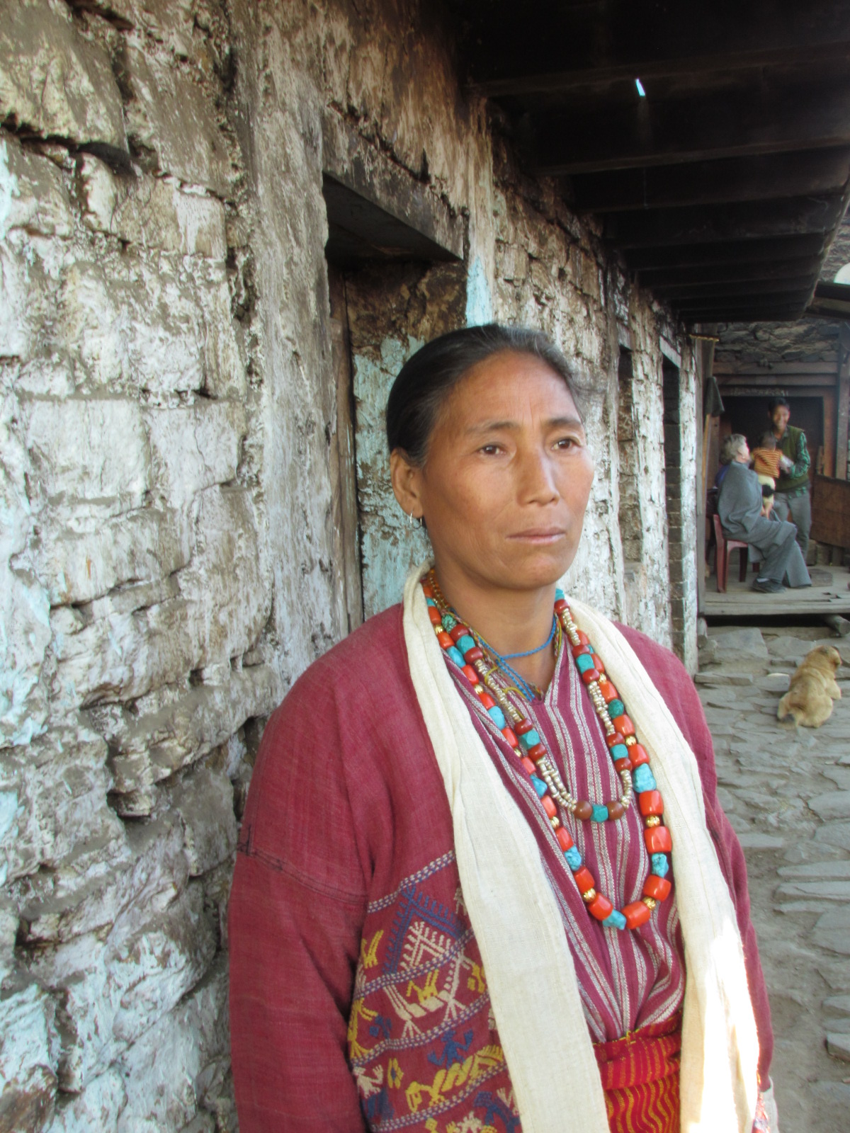 Monpa woman in a hand-loomed striped jacket wearing coral and turquoise necklaces, standing outside a whitewashed stone house in West Kameng, Arunachal Pradesh