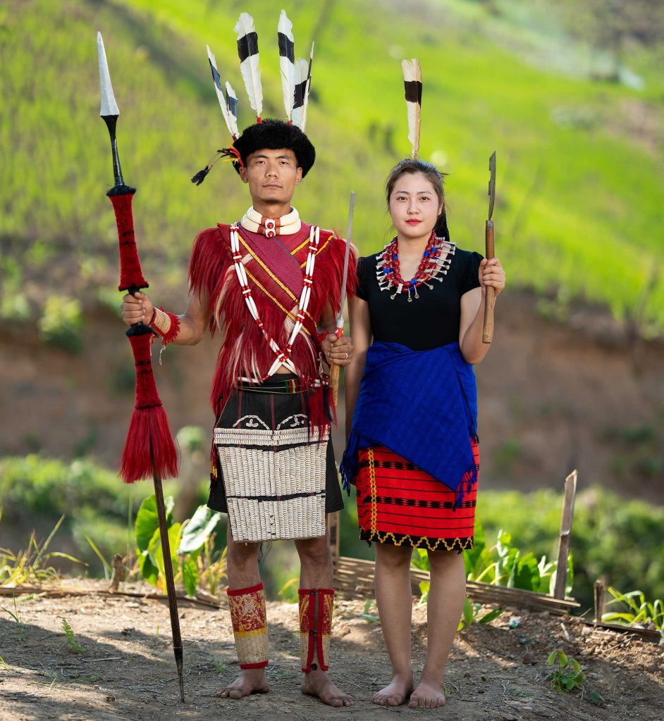 Ao Naga couple in traditional Moatsu Festival attire — feathered headdress, red goat-hair shawl and woven basket, Mokokchung, Nagaland